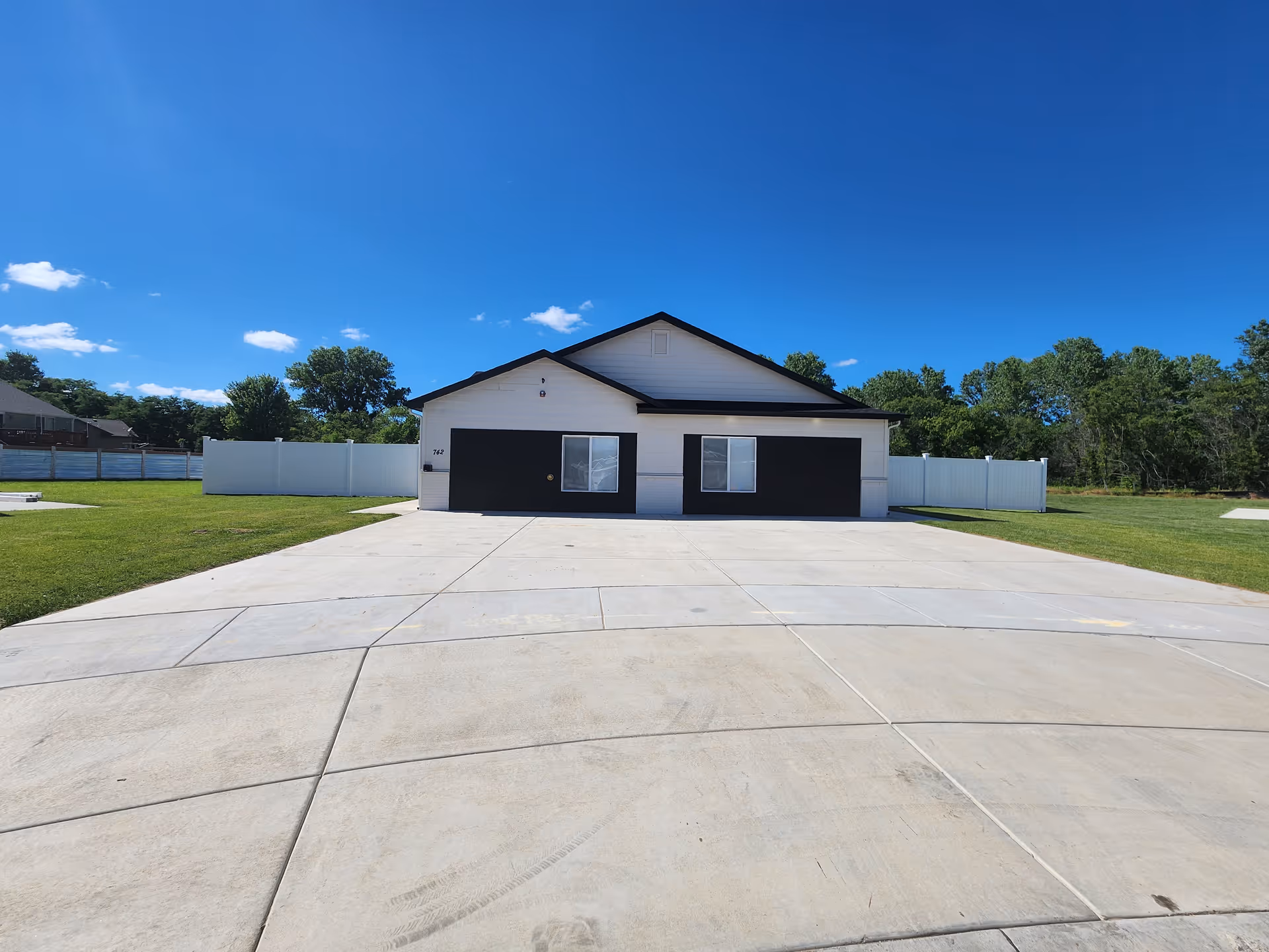 Front exterior view of a single-story building with white siding and black trim, including two black garage doors and two windows. The building is surrounded by a large concrete driveway and green grass lawn with a white fence in the background under a clear blue sky.