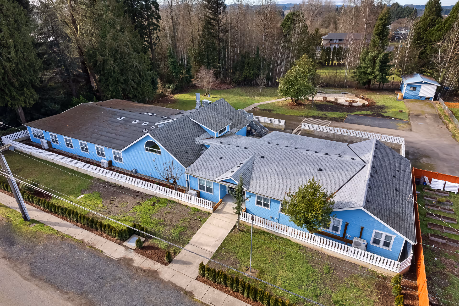 Aerial view of Wellspring Centralia, a single-story blue building with a gray roof surrounded by a white picket fence. The building is situated in a green area with trees and a small garden space. There is a sidewalk leading to the entrance and a small shed in the background.