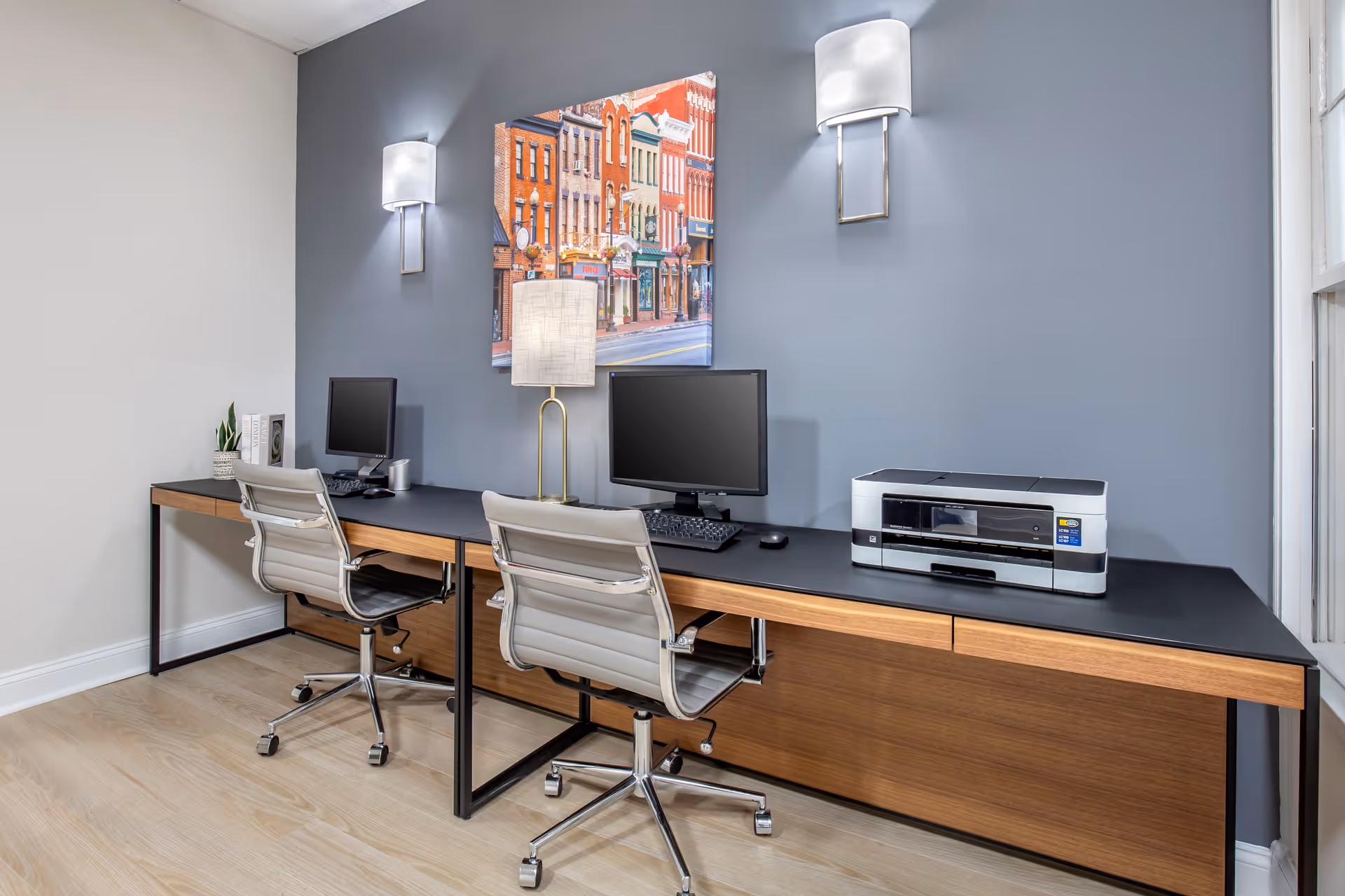 A modern office area with two workstations featuring computers, a printer, and two gray office chairs on wheels. The wall behind the desks is painted blue and has two wall-mounted lights and a colorful painting of a street with buildings. A table lamp is placed between the two computer monitors.
