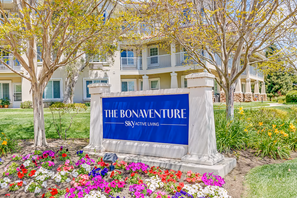 Outdoor view of a senior living facility with a large blue sign reading 'THE BONAVENTURE SKY ACTIVE LIVING' surrounded by colorful flowers and trees, with a multi-story building in the background.