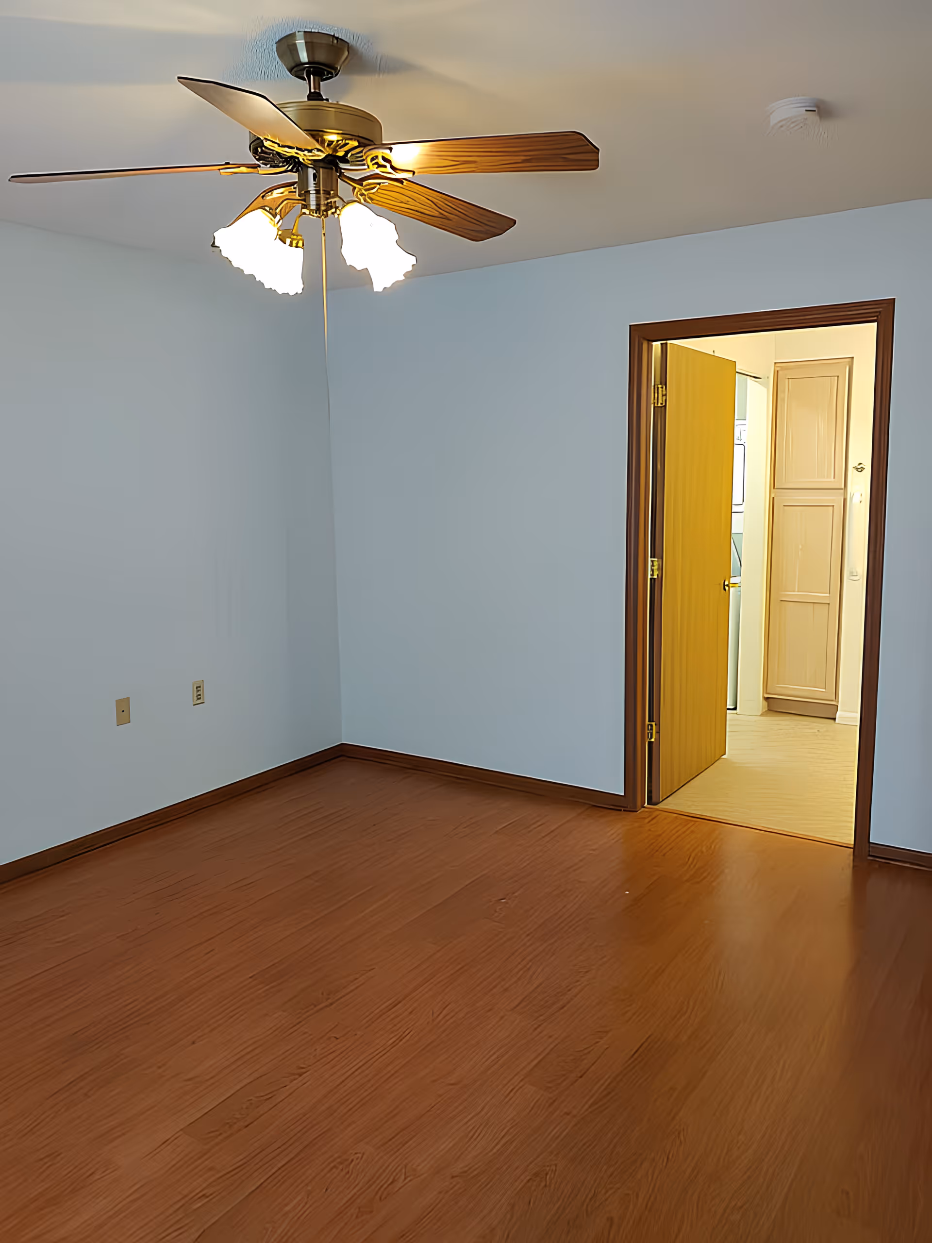 Empty room with wooden floor, light blue walls, a ceiling fan with lights, and an open door leading to another room with cabinets and appliances.