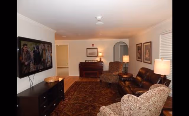 Interior living room with a wall-mounted TV, leather sofa, patterned armchairs, area rug, lamps, and a piano in the background.