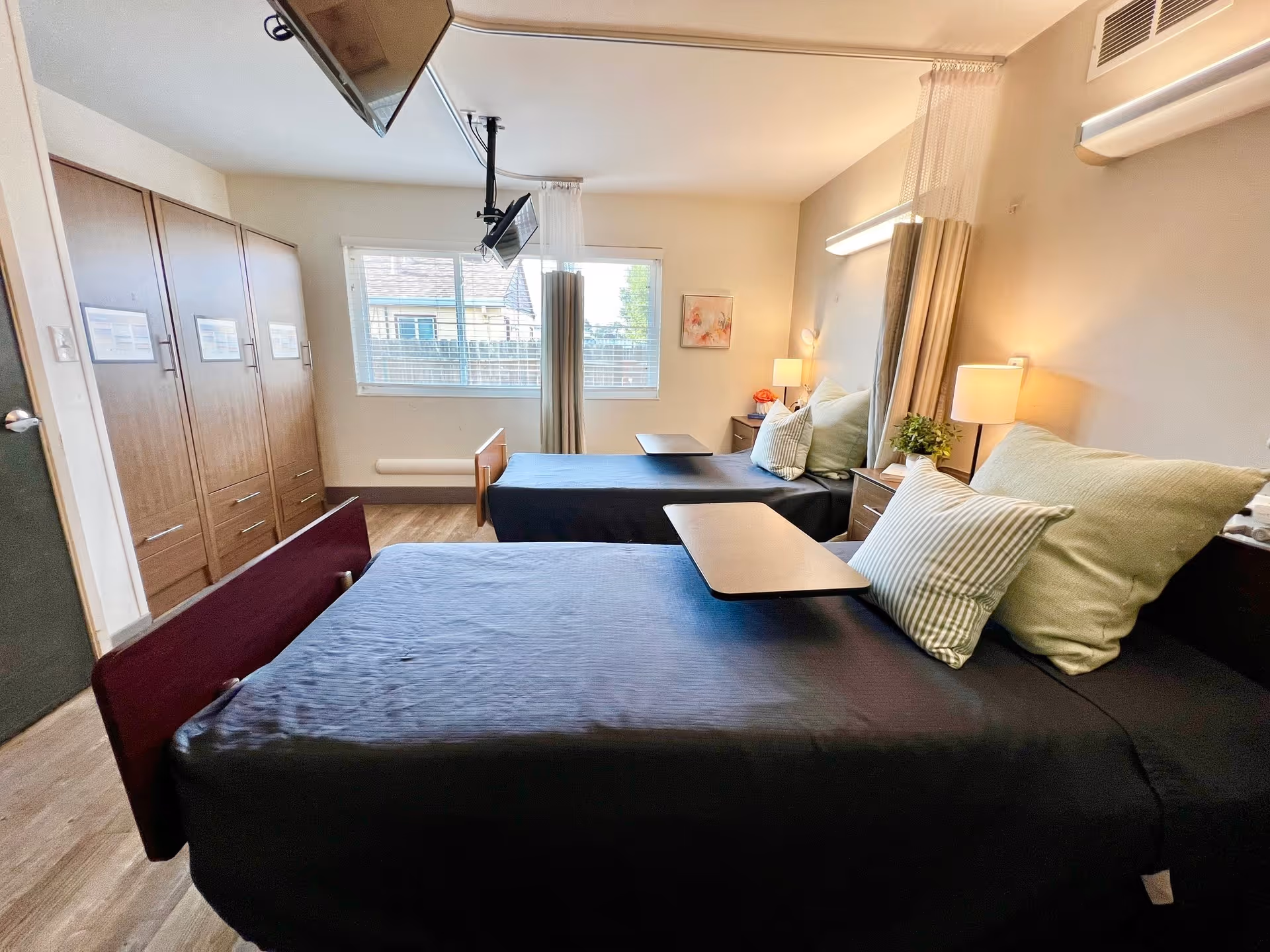 A bright and clean bedroom in a healthcare facility with two hospital-style beds covered in dark bedding, each with pillows and an overbed table. There are wooden wardrobes along one wall, a window with blinds letting in natural light, and bedside tables with lamps and small plants. A ceiling-mounted TV is positioned above the beds.