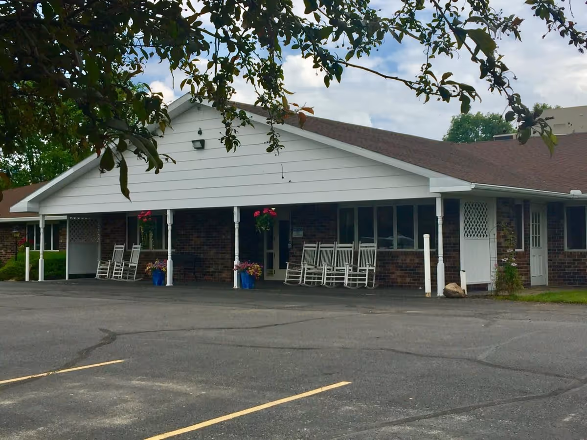 Exterior view of a single-story brick building with a white gabled roof and a covered porch. The porch features several white rocking chairs and hanging flower baskets. The foreground shows an empty paved parking lot with yellow parking lines. Trees with green leaves partially frame the top of the image under a partly cloudy sky.