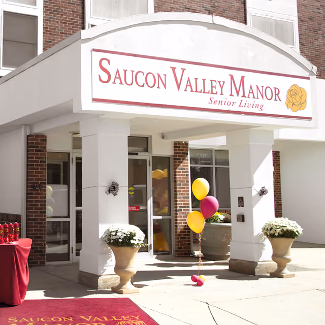 Entrance of Saucon Valley Manor senior living facility with a white and brick exterior, two large white columns, potted plants with white flowers, a red carpet with the facility name, and yellow and purple balloons tied to a weight.