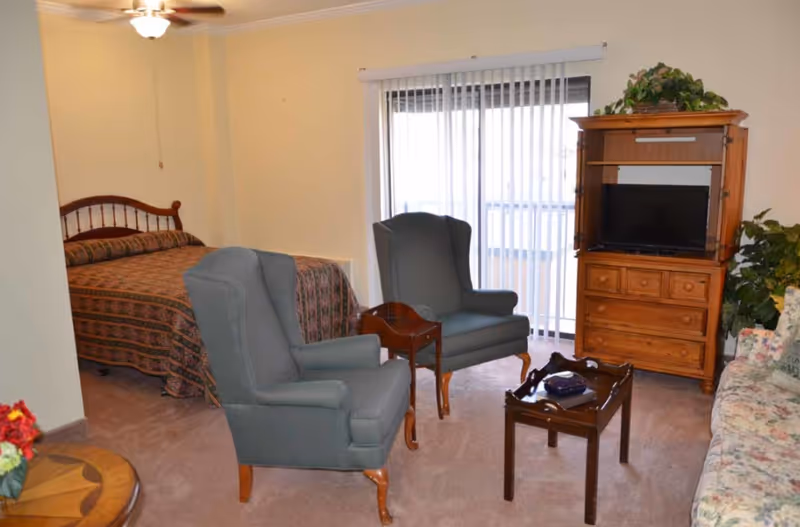 A cozy room in a senior living facility featuring a bed with a patterned bedspread, two upholstered armchairs, a wooden TV cabinet with a television, a small wooden coffee table, a floral-patterned sofa, and a sliding glass door with vertical blinds leading to a balcony or outdoor area.
