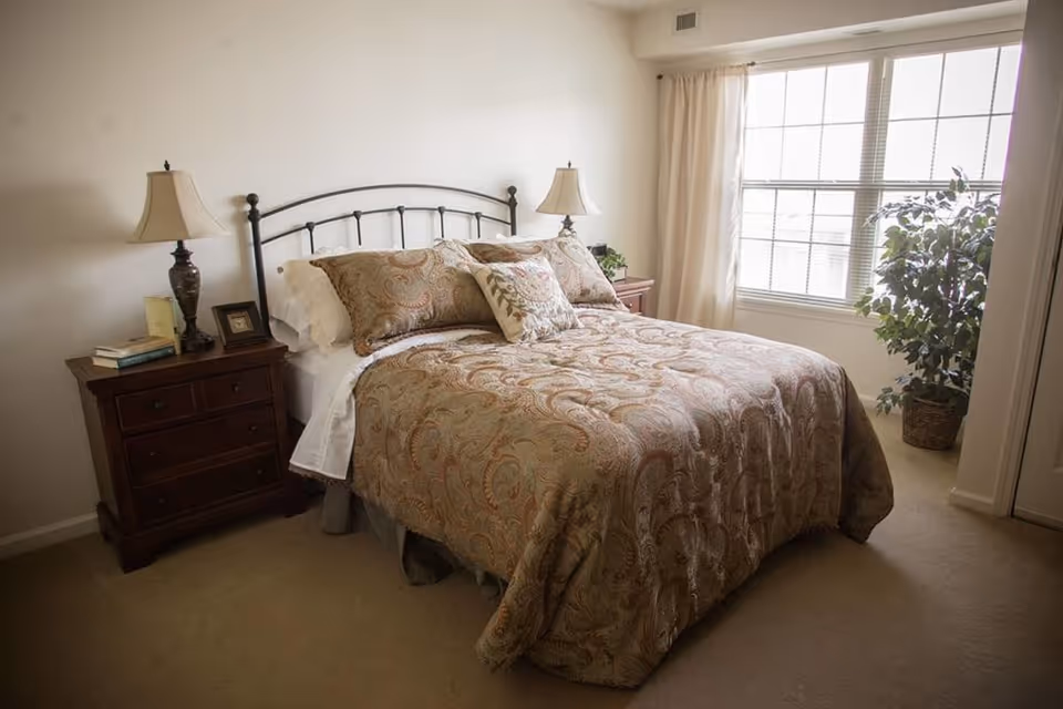 A neatly made bedroom with a patterned bed, metal headboard, two nightstands with lamps, and a window with a potted plant.