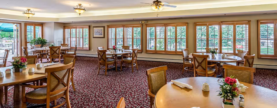 A spacious dining room with multiple round and rectangular wooden tables and chairs. Each table is set with napkins, cups, and small flower arrangements. Large windows with wooden shutters allow natural light to fill the room, and a ceiling fan and light fixtures are visible on the ceiling. The carpet has a red and white patterned design.
