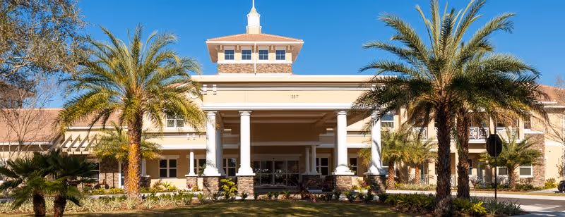 Front exterior view of HarborChase of Villages Crossing building with a covered entrance supported by white columns, surrounded by palm trees and landscaping under a clear blue sky.