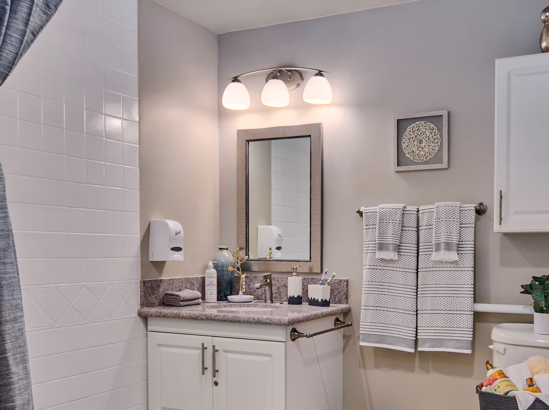 A clean and modern bathroom featuring a white tiled shower with a gray curtain, a granite countertop with a sink, a mirror with three overhead lights, neatly folded towels on a towel rack, and various toiletries on the counter. There is also a white cabinet and a basket with rolled towels and other items near the toilet.