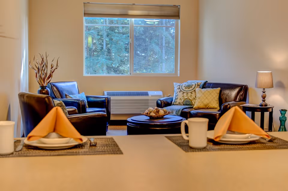 A cozy living room area with two dark leather armchairs and a matching loveseat adorned with decorative pillows. A round coffee table with a decorative bowl is centered in front of the seating. A window behind the seating area shows a view of trees outside. In the foreground, a dining counter is set with two place settings, each featuring a folded orange napkin, a plate, and a white mug. A table lamp and decorative vases are visible on side tables.