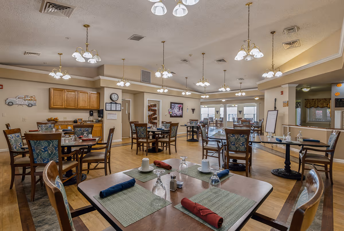A spacious dining room in a senior living facility with multiple wooden tables and chairs arranged neatly. Each table is set with placemats, napkins, cups, and glasses. The room is well-lit with several hanging light fixtures and has a wooden floor. There are cabinets and a clock on one wall, and large windows in the background letting in natural light.