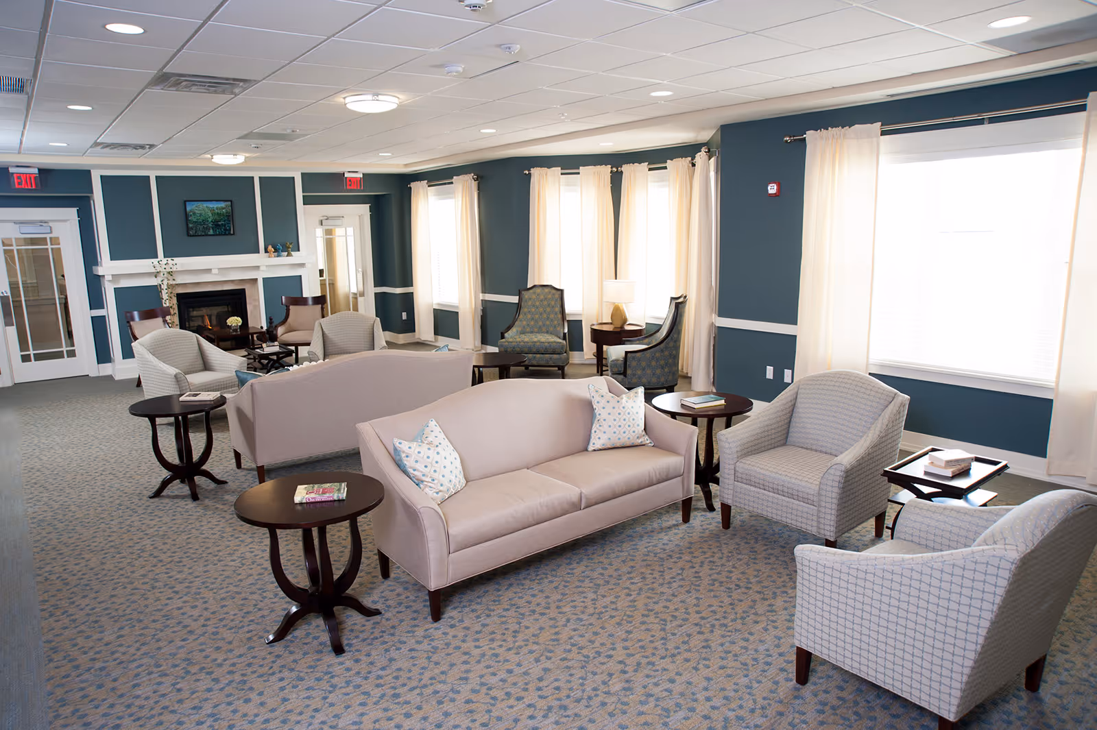 A spacious and well-lit living room with multiple seating arrangements including sofas and armchairs. The room features large windows with sheer curtains, a fireplace with a mantel, side tables with books, and soft carpeting. The walls are painted dark teal with white trim, and there are overhead lights in the ceiling.