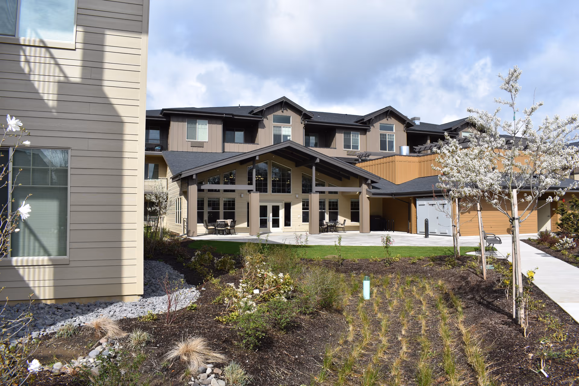 Exterior view of a multi-story senior living facility building with beige and brown siding, large windows, and a covered patio area with tables and chairs. The foreground features landscaped garden beds with small plants and flowering trees under a partly cloudy sky.