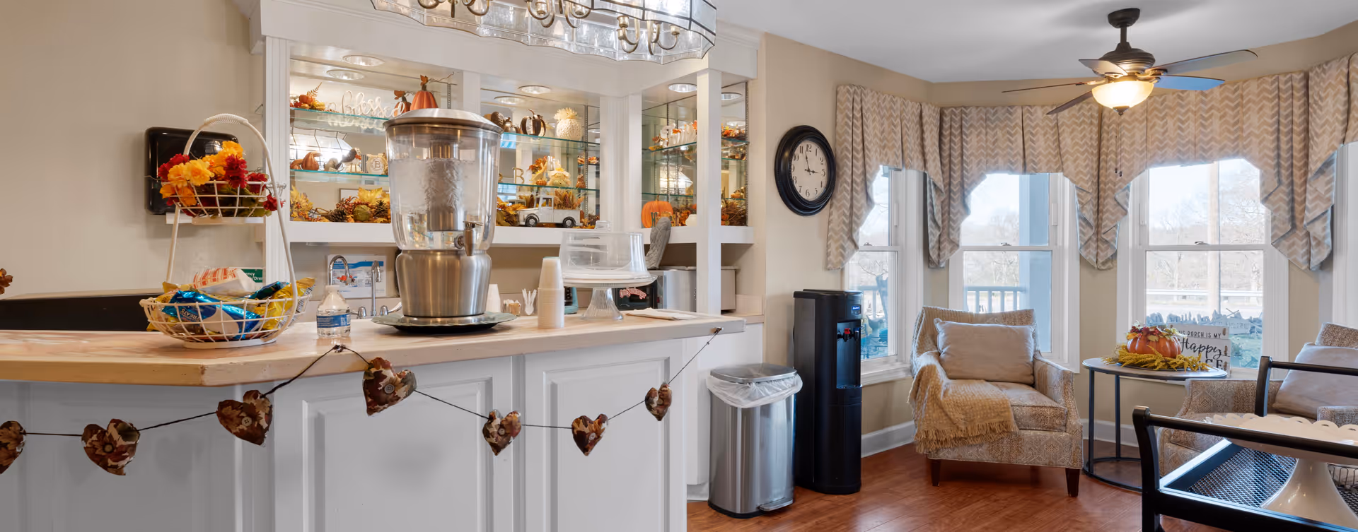 A cozy interior space featuring a white counter with a water dispenser and fall-themed decorations, including a garland of leaves. Behind the counter is a mirrored wall with shelves displaying seasonal decor. To the right, there are two cushioned armchairs near large windows with patterned valances, a small round table with a fall centerpiece, and a ceiling fan with a light fixture.