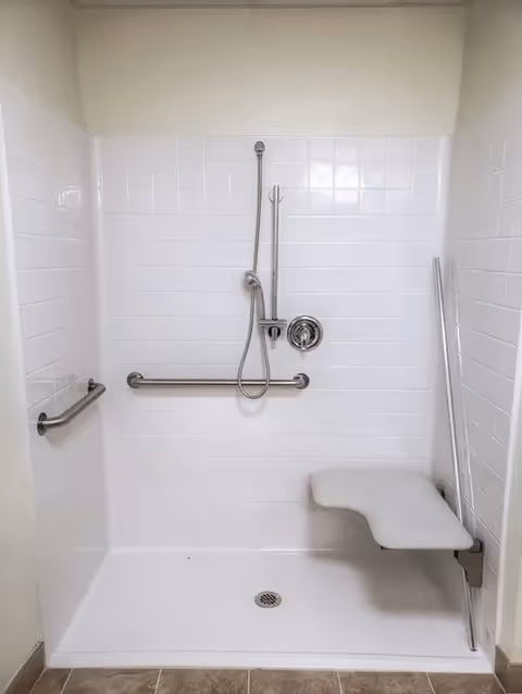 A white tiled walk-in shower with a handheld showerhead attached to a vertical sliding bar, a horizontal grab bar on the back wall, and a fold-down shower seat on the right side. The floor is white with a central drain, and the surrounding floor outside the shower is tiled in a beige color.