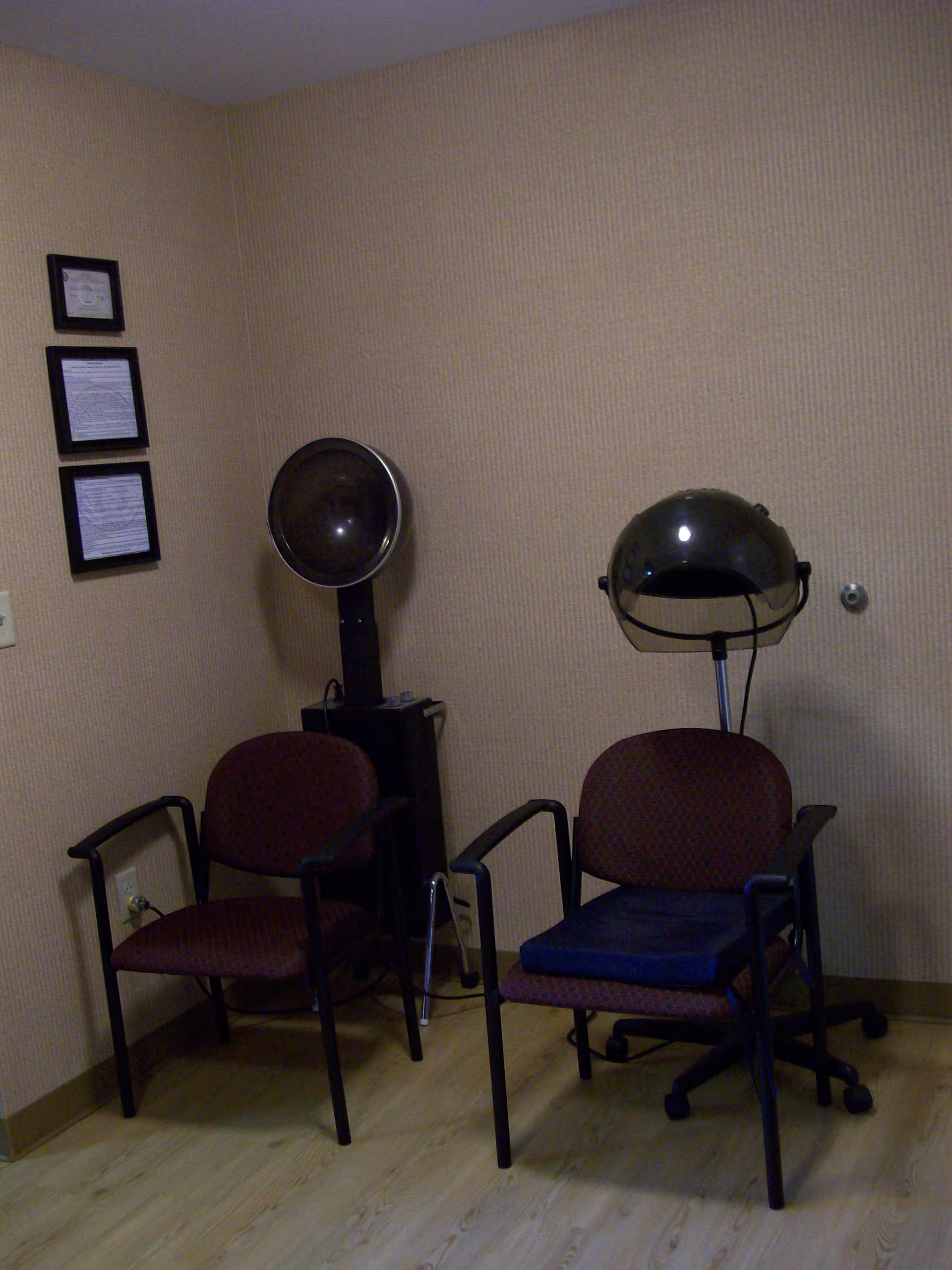 A small room with two maroon chairs, one with a blue cushion, and two vintage hair drying machines. The walls are beige with a subtle pattern, and there are three framed documents hanging on the left wall. The floor is light wood.