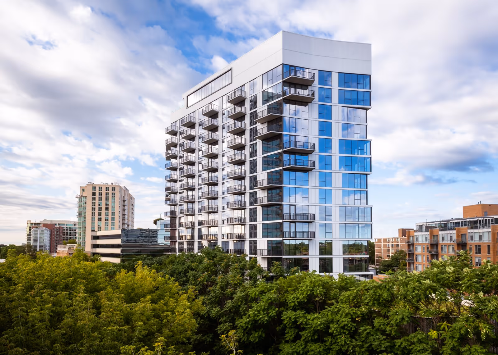 Modern multi-story apartment building with numerous balconies, surrounded by trees and other buildings under a partly cloudy sky.
