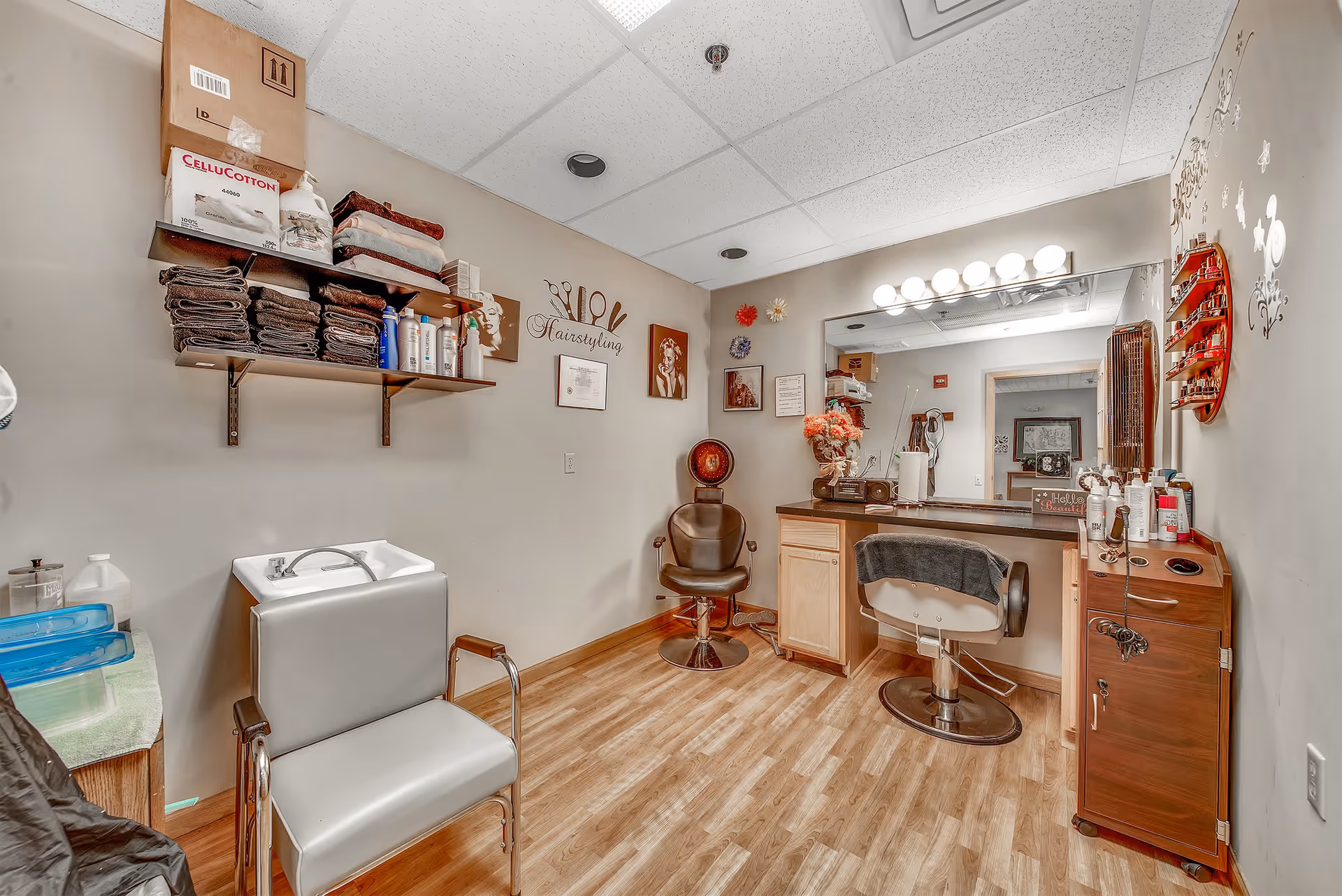 Interior view of a hairstyling room in a senior living facility. The room features a styling chair, a sink for washing hair, shelves stocked with towels and hair care products, a large mirror with lights above it, and various decorative items on the walls.