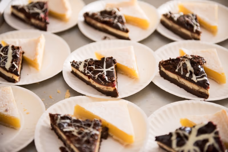 Multiple white paper plates each holding two slices of dessert: one slice of chocolate cake with white icing drizzle and one slice of lemon tart with powdered sugar on top.