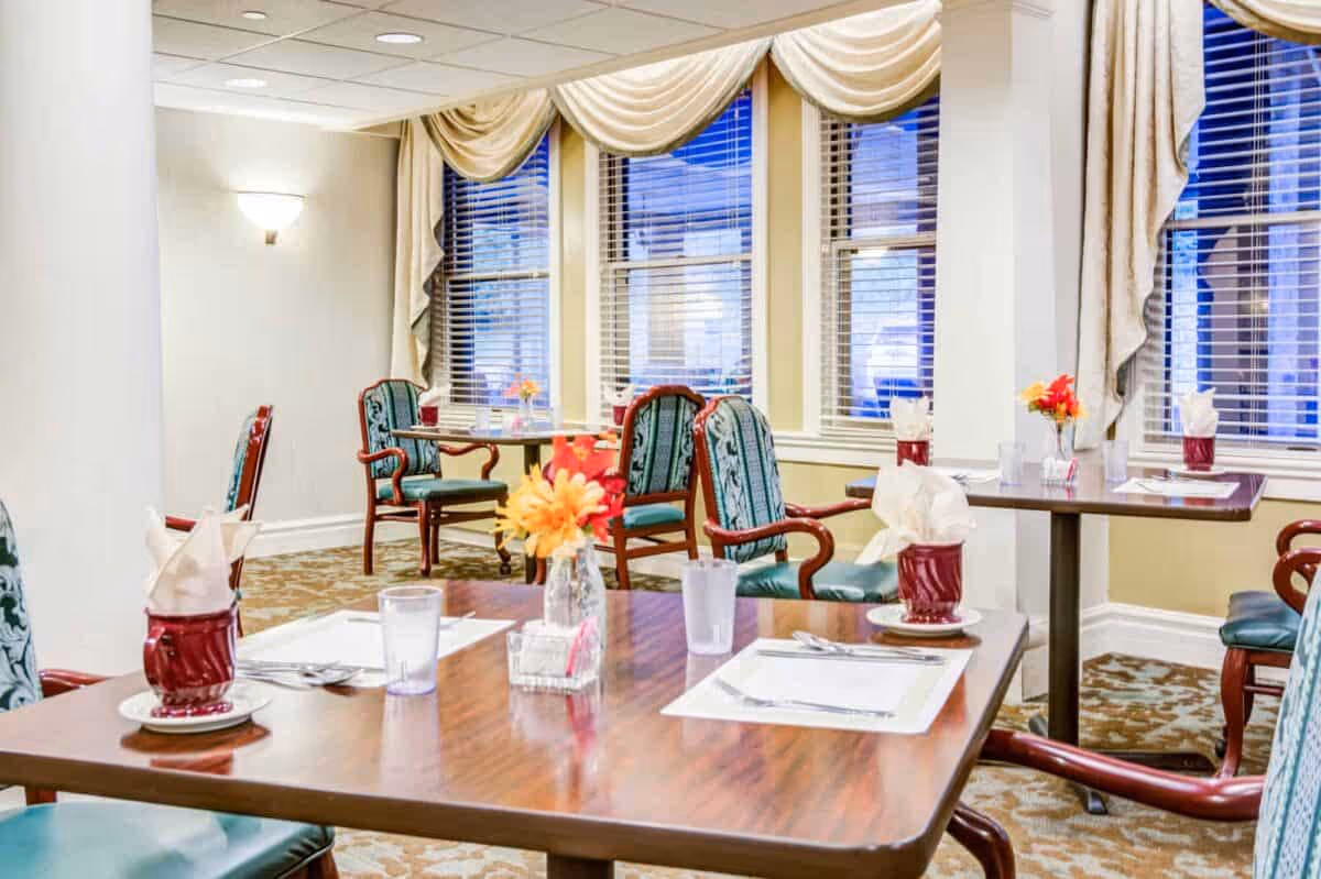 Dining room with multiple set tables and upholstered chairs, floral centerpieces, and large windows with blinds and draped valances.
