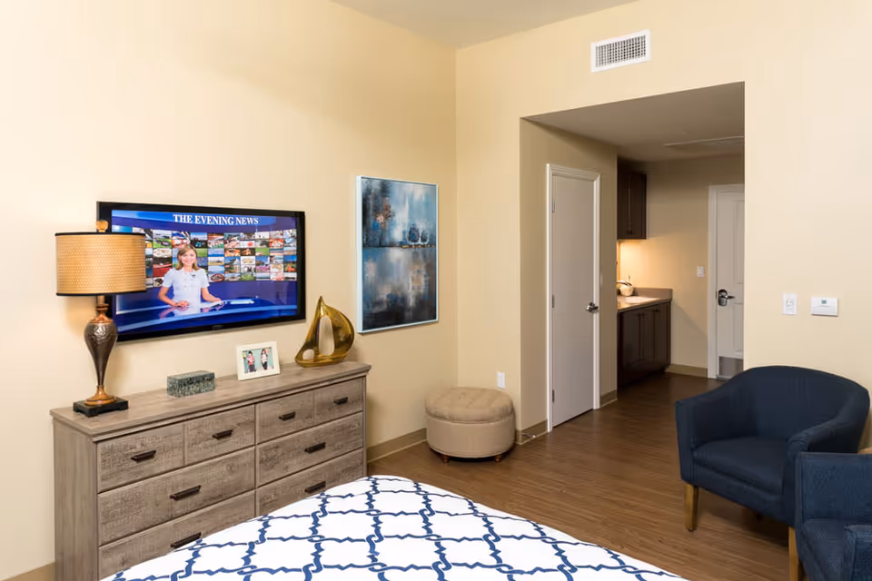 A cozy bedroom in a senior living facility with a bed covered in a white and blue patterned blanket. A wooden dresser with a lamp, decorative items, and a framed photo sits against the wall beneath a mounted flat-screen TV displaying a news program. There is a blue armchair, a small round ottoman, and an open doorway leading to a kitchenette area with cabinets and a countertop.