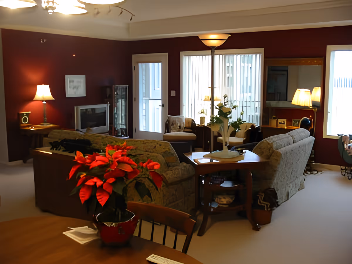 A cozy living room with maroon walls, two patterned sofas facing each other, a wooden coffee table with a white vase and flowers, a floor lamp, and a side table with a lamp. There is a TV on a stand against the wall, a glass display cabinet, and large windows with vertical blinds letting in natural light. A red poinsettia plant is on a wooden dining table in the foreground.