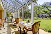 A bright sunroom dining area with glass walls and ceiling overlooking a green garden. Several tables are set with white tablecloths, plates, and utensils, each accompanied by cushioned wooden chairs. The outdoor garden features trees and a well-maintained lawn.