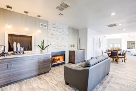 Bright communal living area with a sofa facing a stone fireplace, built-in cabinets, and a dining table visible in the background.