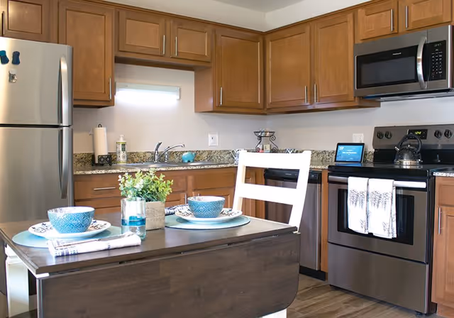 A modern kitchen with wooden cabinets, stainless steel appliances including a refrigerator, microwave, stove, and dishwasher. A small dining table with two place settings, blue bowls, plates, and napkins is in the foreground. A small plant and a glass of water are on the table. The kitchen has granite countertops and a white wall with a small window above the sink.