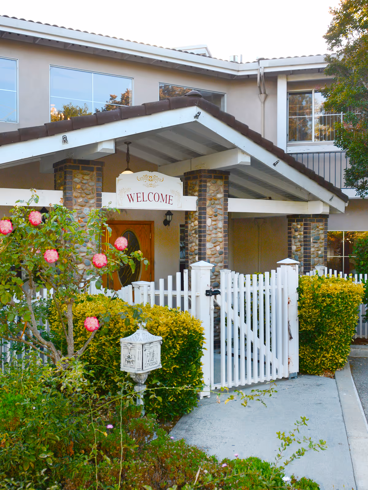Entrance of a senior living facility with a white picket fence gate, stone pillars supporting a covered porch, a wooden door, and a sign that reads 'WELCOME'. There are bushes and pink flowers in the garden area near the entrance.