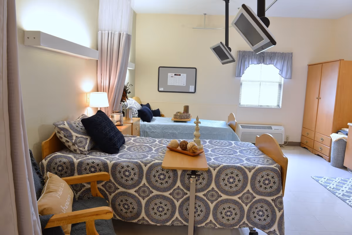 A bright shared bedroom with two twin beds, patterned bedspreads, a bedside lamp, mounted televisions, and wooden storage furniture.