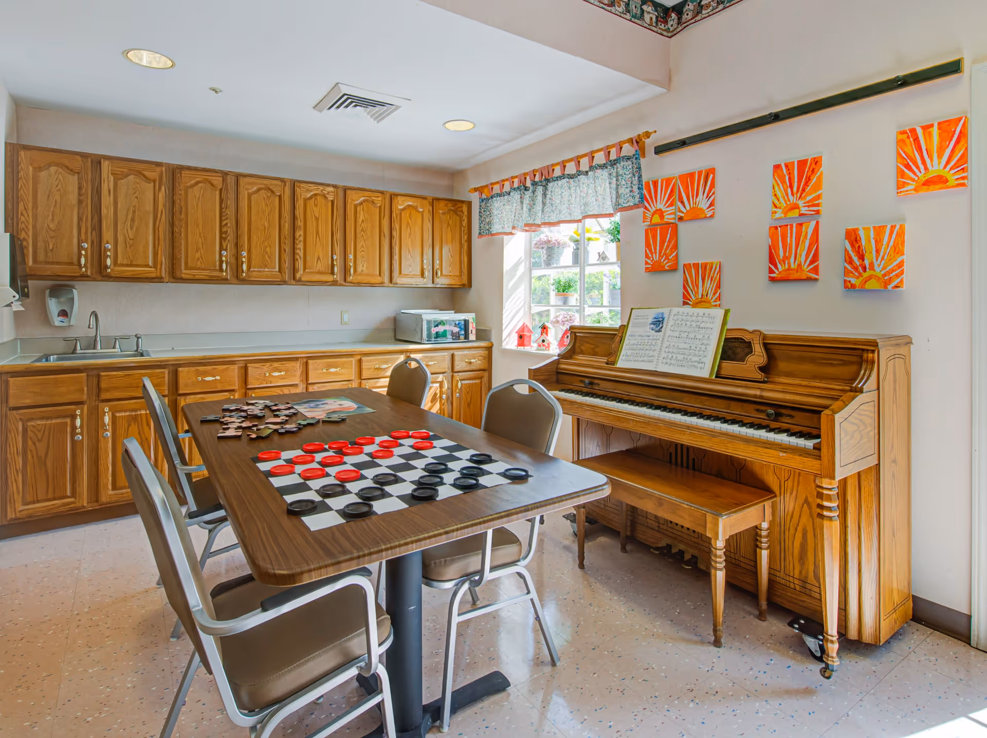 A bright room with wooden cabinets along one wall, a table with chairs around it featuring a checkers game and a puzzle, and an upright piano with sheet music on it. The wall above the piano is decorated with colorful paintings of suns, and a window with a floral valance lets in natural light.