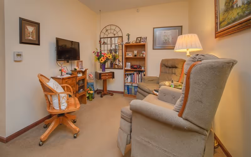 A cozy living room area with two cushioned armchairs, a wooden swivel chair with a white pillow, a small wooden cabinet with a flat-screen TV mounted above it, a bookshelf filled with books and decorative items, a side table with a vase of flowers, and a floor lamp providing warm light. The walls are adorned with framed artwork and a decorative mirror.