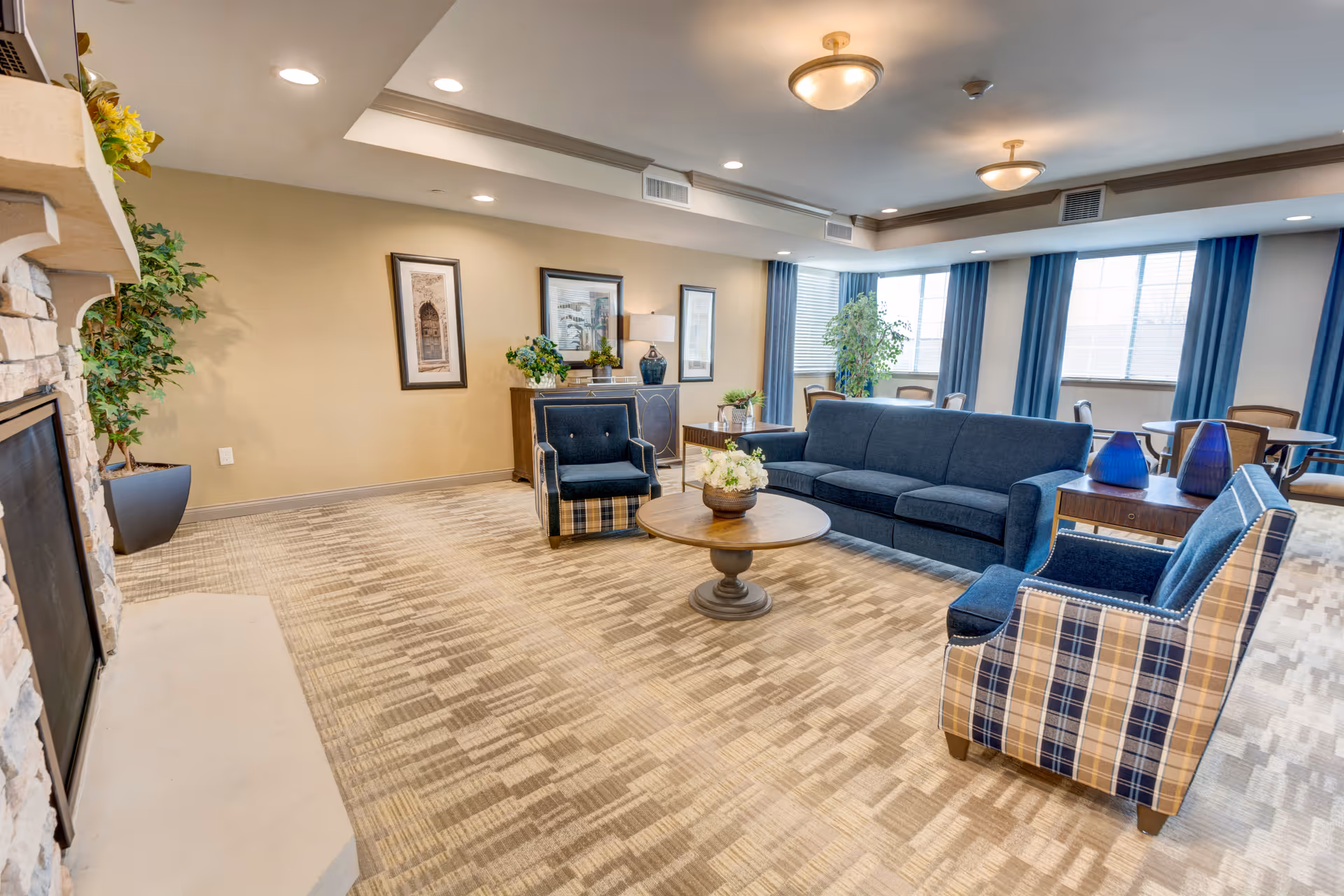 A spacious living room area with a blue sofa, two plaid armchairs, a round wooden coffee table with a flower arrangement, and a stone fireplace on the left. The room has beige walls, framed artwork, potted plants, and large windows with blue curtains letting in natural light. There are also ceiling lights and a side table with decorative blue vases.