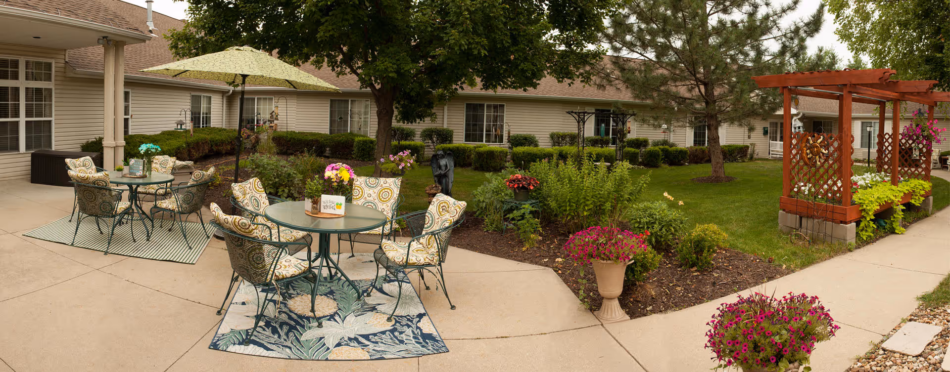 Outdoor patio area at Bickford of Cedar Falls with round metal tables and cushioned chairs on outdoor rugs, surrounded by flower pots and garden beds with various plants and flowers. There is a large umbrella providing shade over one table, and a wooden pergola with hanging flower baskets on the right side. The building exterior with windows and beige siding is visible in the background.