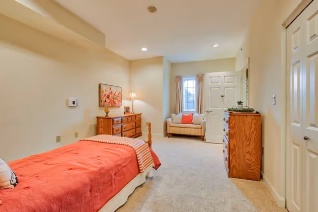 A cozy bedroom with a single bed covered in a red bedspread and a striped throw blanket. There is a wooden dresser with a lamp and framed picture on top, a floral painting on the wall, and a small cushioned bench with pillows near a window. The room has beige walls and carpeted floor, with a door and closet visible.