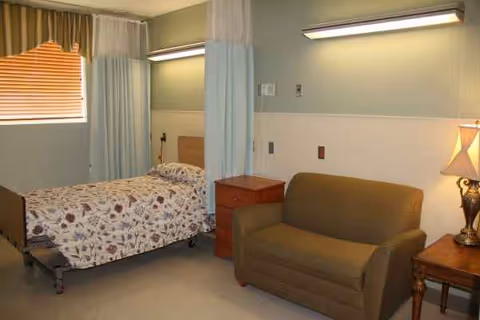 A room with a hospital-style bed covered with a floral patterned blanket, a brown loveseat, a wooden nightstand with a lamp, and a window with blinds and curtains. The room has a privacy curtain partially drawn around the bed.