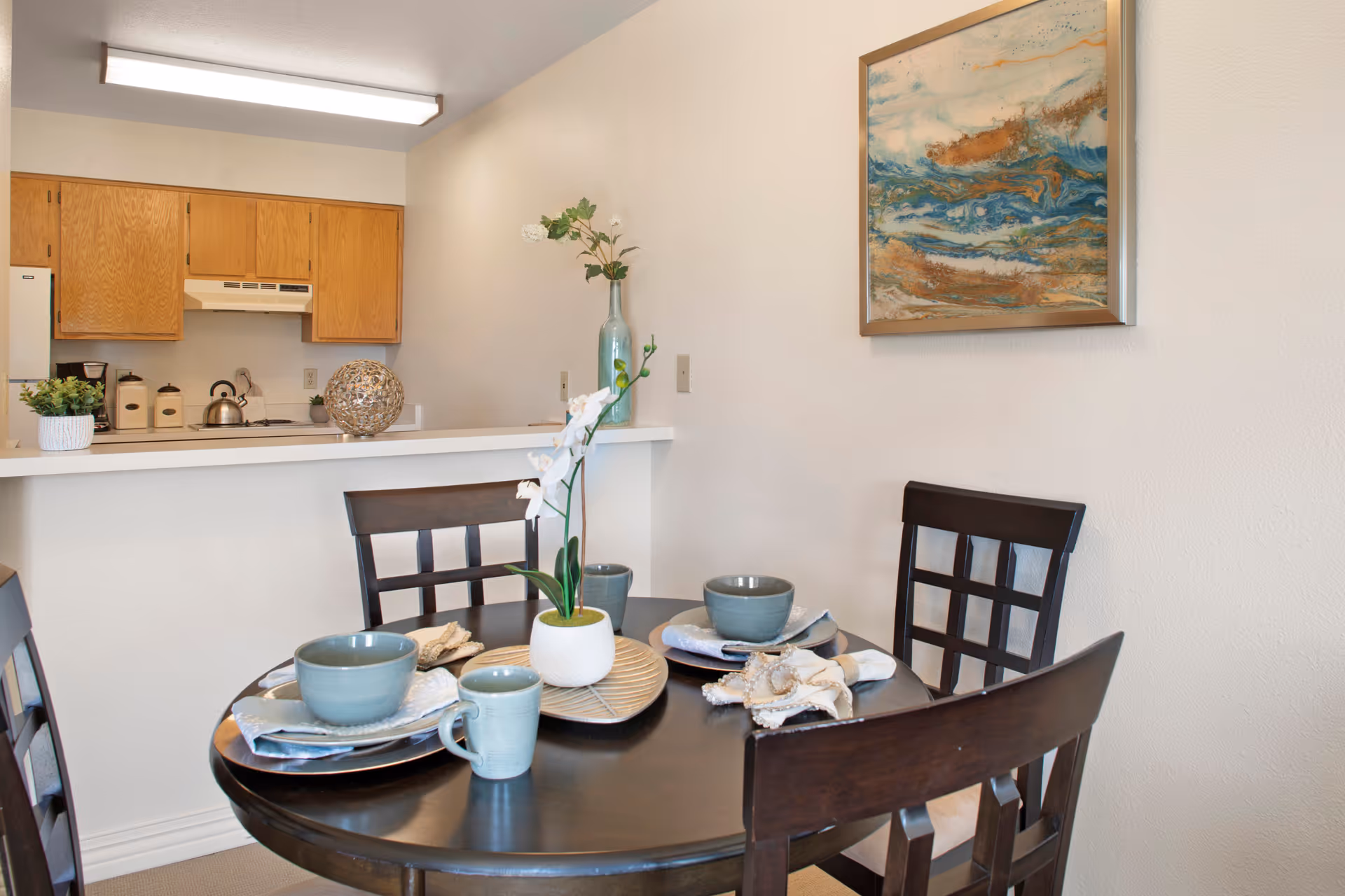 A dining area with a round dark wooden table set for three with blue-gray bowls, plates, mugs, and napkins. A white orchid centerpiece is on the table. Behind the table is a kitchen with wooden cabinets, a white countertop, and a kettle on the stove. A framed abstract painting with blue, white, and brown tones hangs on the wall.