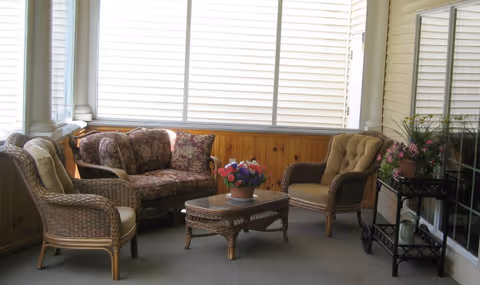 A sunroom with wicker furniture including a loveseat with floral cushions, two armchairs with beige cushions, and a wicker coffee table with a flower pot on top. The room has large windows with closed blinds, wood paneling on the lower walls, and a small black metal cart with plants and decorative items.
