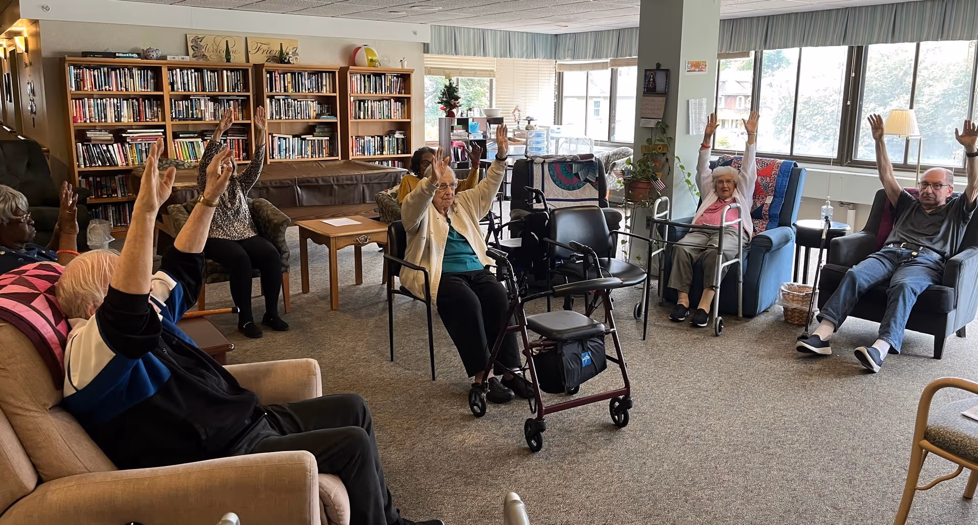 Seniors seated in a communal living room raising their arms during a group exercise, with bookshelves and large windows in the background.