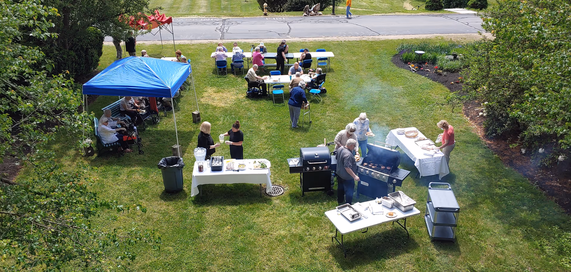 A group of elderly people and caregivers enjoying an outdoor barbecue event on a grassy lawn. Several tables with food and drinks are set up, with two grills in use. Some people are seated under a blue canopy, while others are sitting at tables or standing around the grills. Trees and a road are visible in the background.