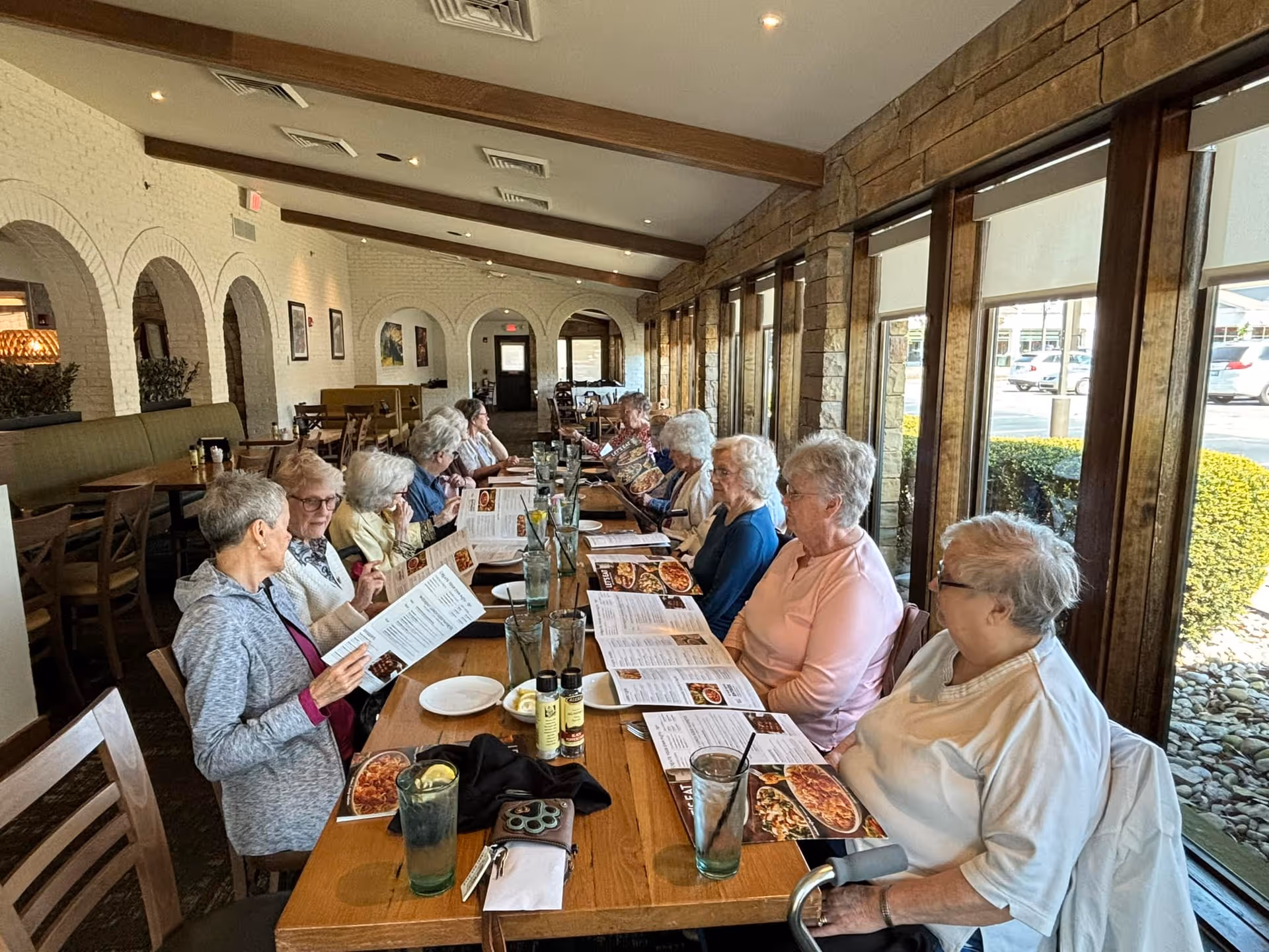 A group of elderly women sitting at a long wooden table in a restaurant dining area, looking at menus. The room has large windows on one side with a view of the parking lot outside, and white brick walls with arches on the opposite side.