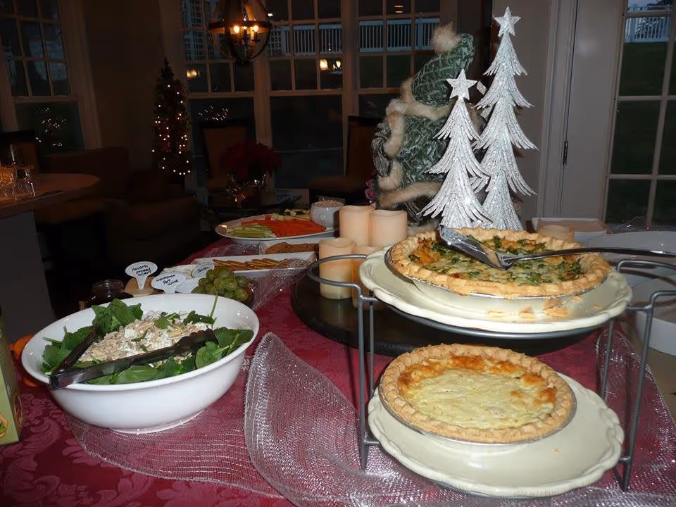 A festive buffet table set with two quiches on a tiered stand, a bowl of salad with tongs, a plate of cut vegetables, grapes, and several candles. In the background, there are Christmas decorations including two white decorative trees and a small Christmas tree with lights near a window.