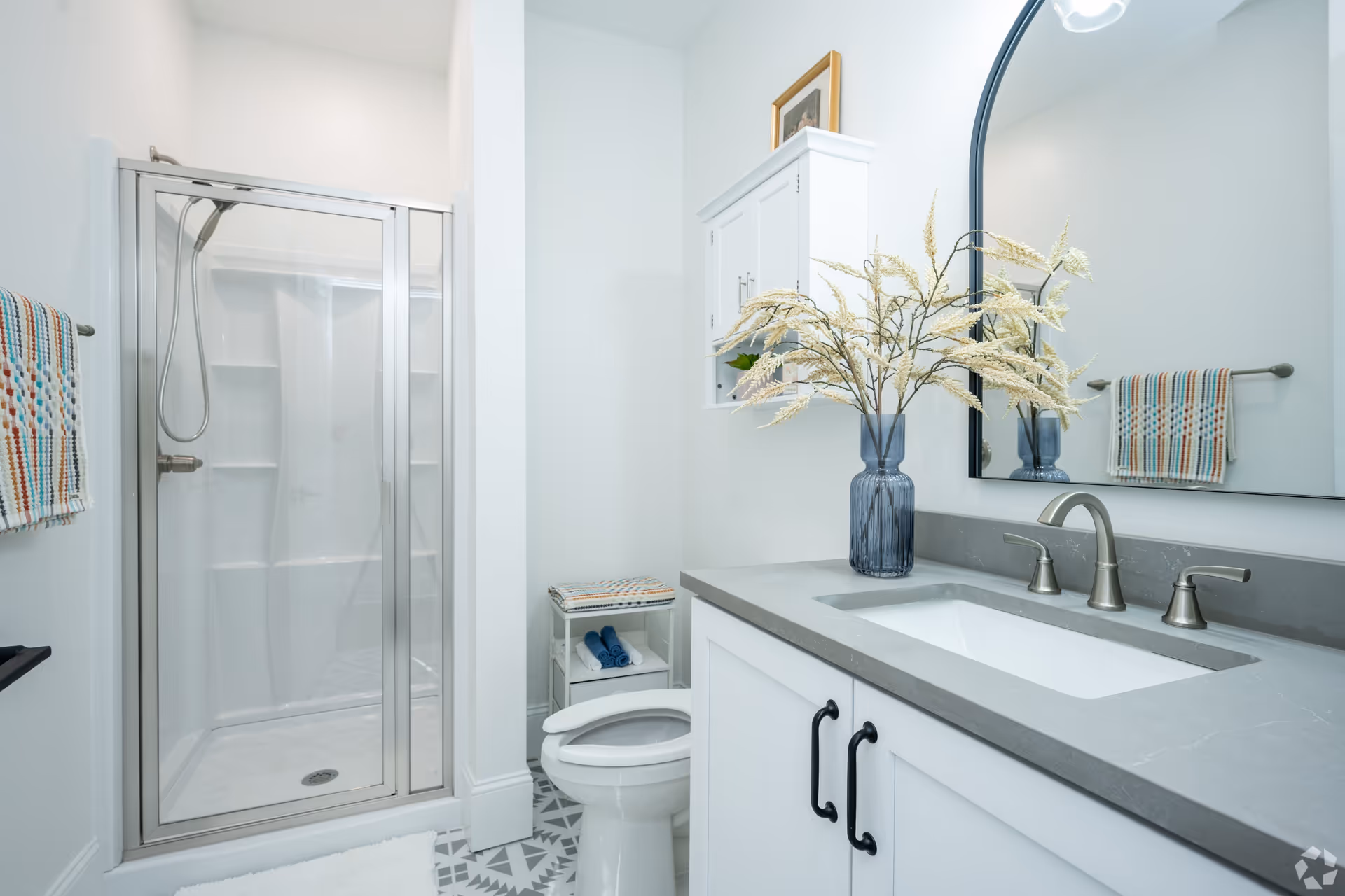 A clean, modern bathroom featuring a glass-enclosed shower with a handheld showerhead, a white toilet, a white vanity with a gray countertop and a built-in sink, a large arched mirror, and a blue vase with decorative dried flowers. There is a small shelf unit next to the toilet holding folded towels, and a colorful striped towel hanging on a towel rack.