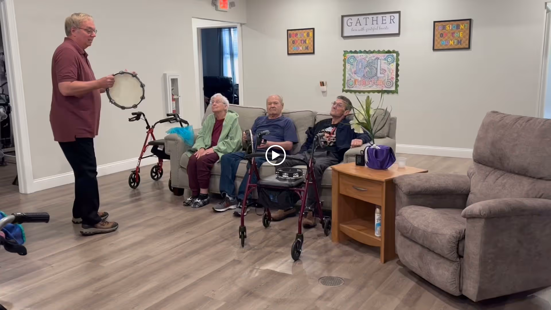 A man holding a tambourine stands in a room with three elderly people seated on a couch. Two walkers are visible near the seated individuals. The room has wood flooring, a side table with a plant and a cup, and wall decorations including a sign that says 'GATHER' and colorful framed artwork.