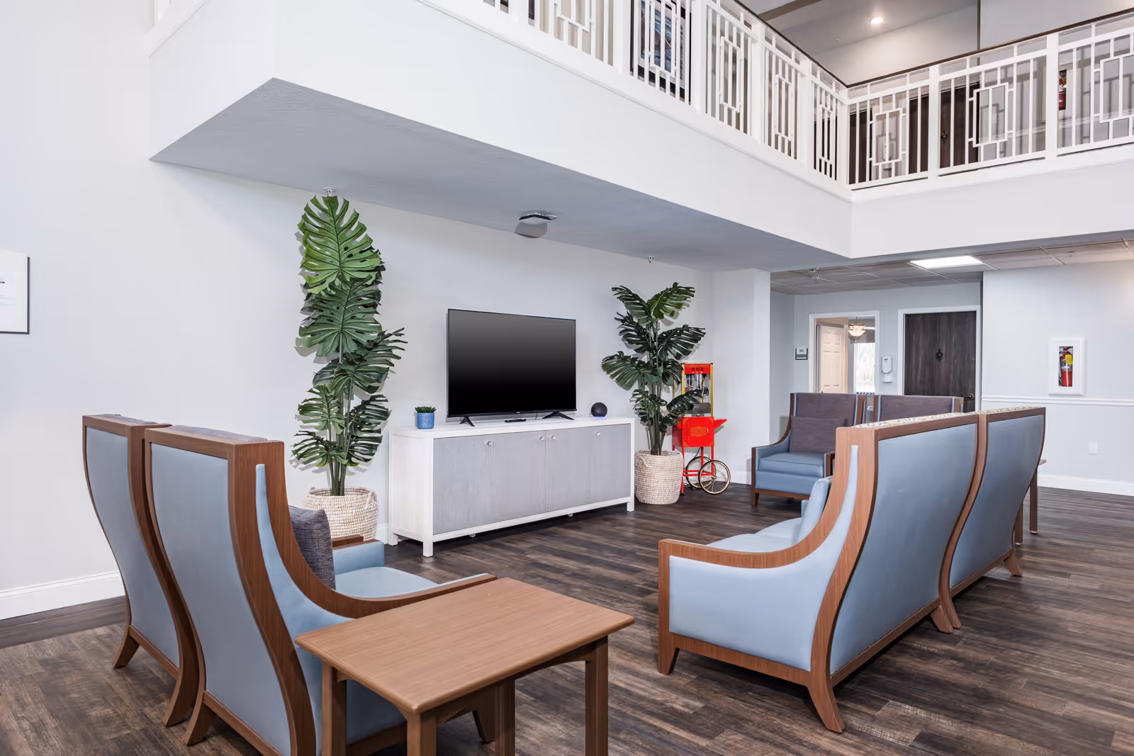 Bright communal living area with a TV, blue-upholstered seating, potted plants, and a small red popcorn cart beneath an open upper balcony.