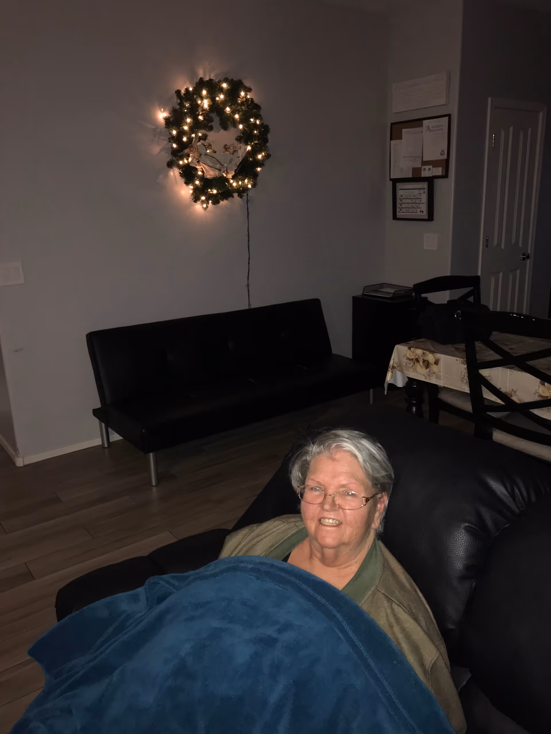An elderly woman with glasses is sitting on a black recliner chair covered with a blue blanket in a dimly lit living room. Behind her, there is a black couch against the wall and a dining table with chairs. A lit Christmas wreath is hanging on the wall above the couch.