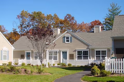 Single-story residential building with beige siding and white trim, featuring multiple windows and a white picket fence in front. A paved walkway leads to the entrance, surrounded by green grass and small shrubs. Trees with autumn-colored leaves are visible in the background under a clear blue sky.