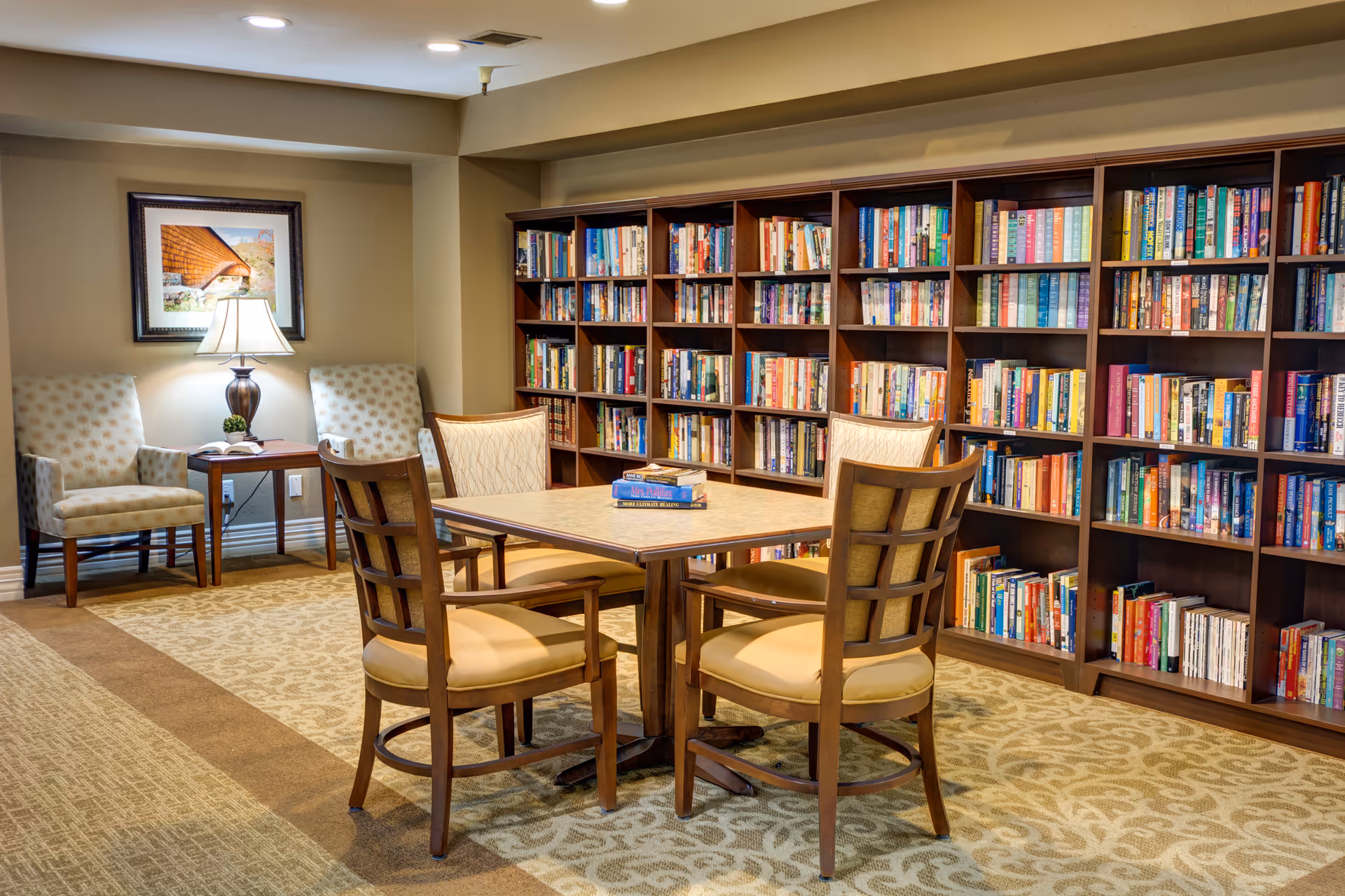 A cozy library room with a large wooden bookshelf filled with books along the wall. In the center, there is a square table with four wooden chairs around it, and a few books placed on the table. In the corner, there are two upholstered armchairs with a small wooden side table between them, topped with a lamp and a framed picture hanging on the wall above.